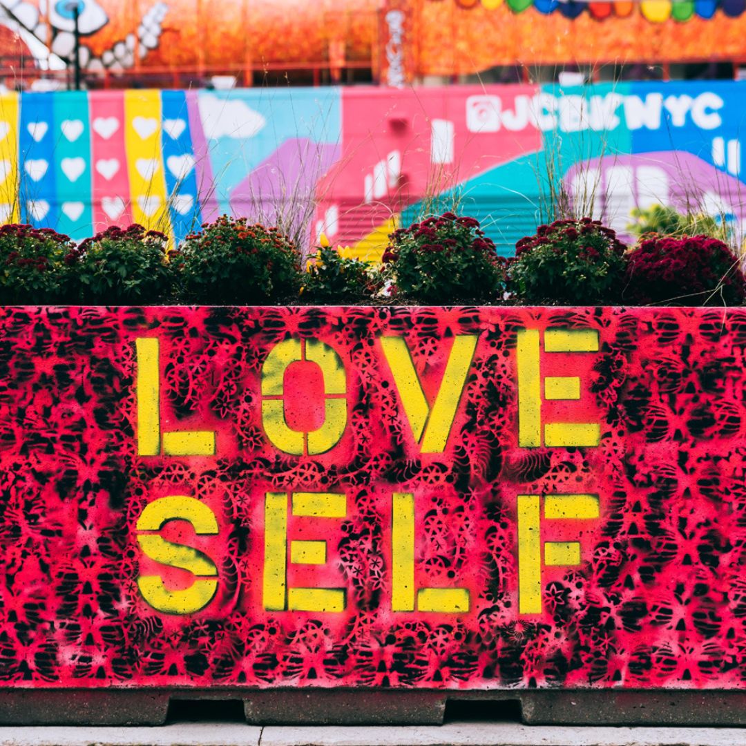 A beautifully coloured creative backdrop with a wall, flowers and the words love self, in yellow on the red and black wall.