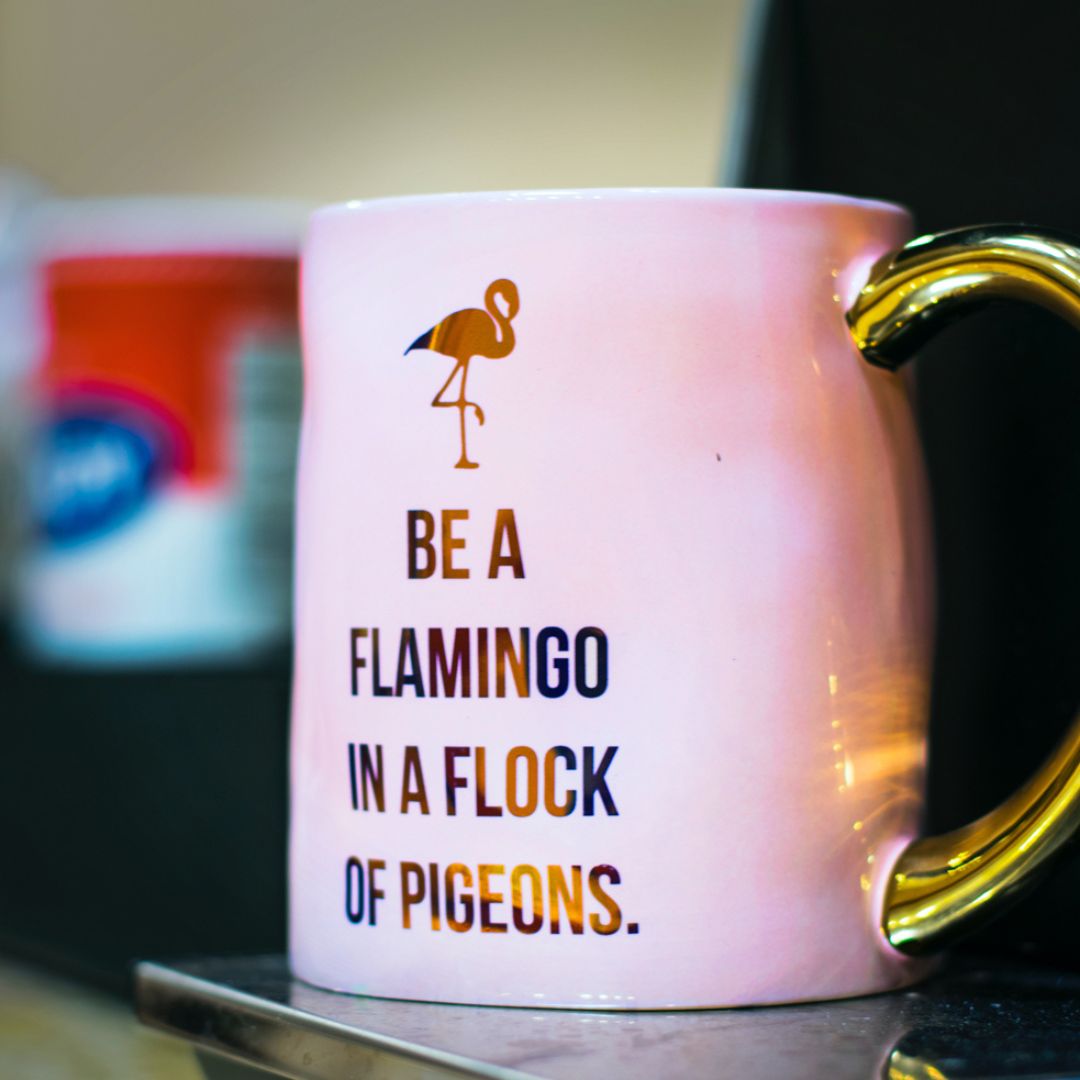A pink cup with a gold handle and a picture of a flamingo with the words be a flamingo in a flock of pigeons.