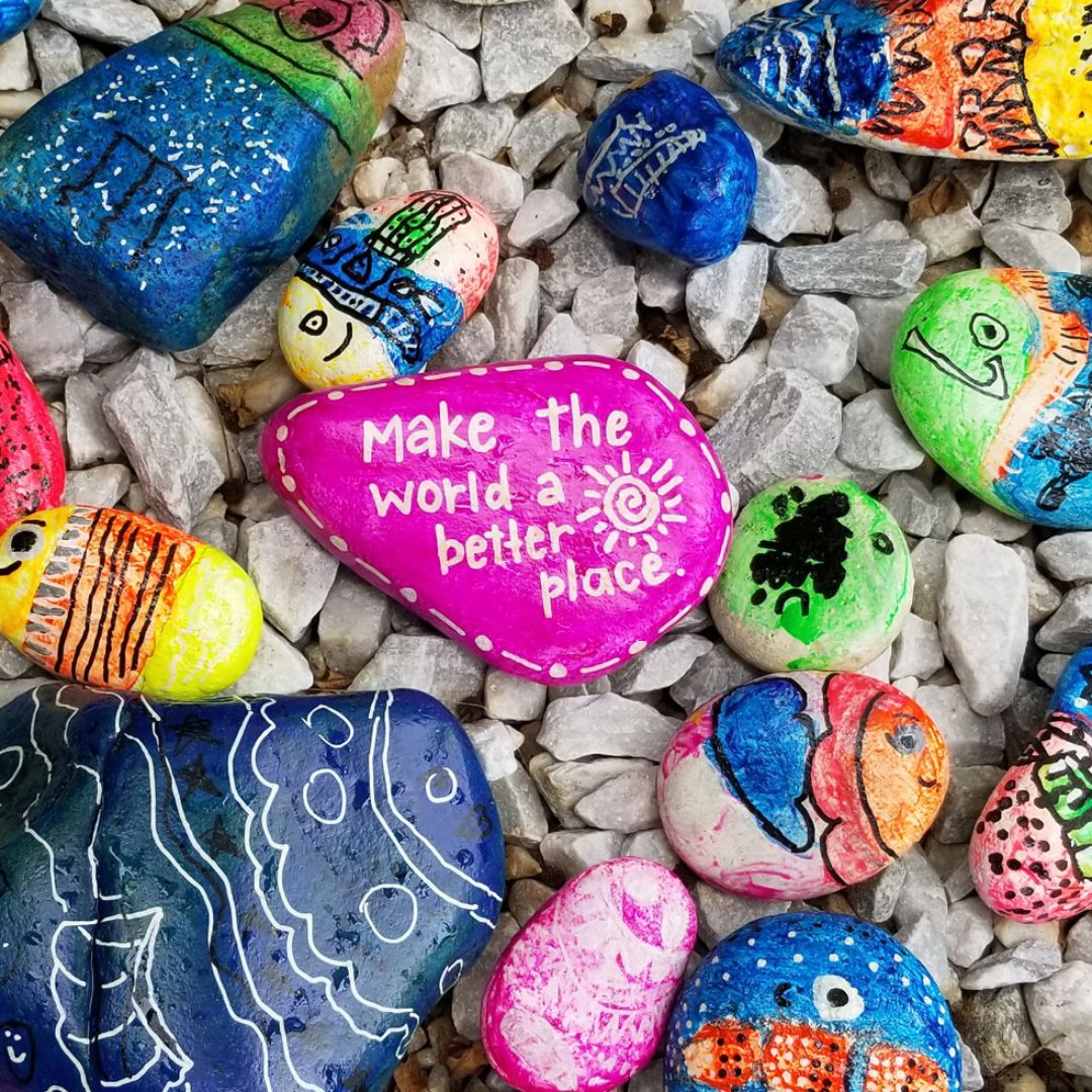 Lots of funky painted pebbles of various sizes and colours with the pink pebble in the middle having the words, make the world a better place.