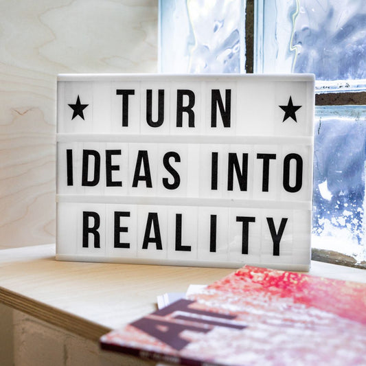 A white plastic board sitting inside a windowsill with some magazines and the words, turn ideas into reality, in black on the white board.