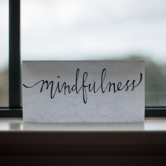 A picture of a window and a piece of paper with the word mindfulness, written on the white paper with black ink and all lit up via the daylight coming through the window.